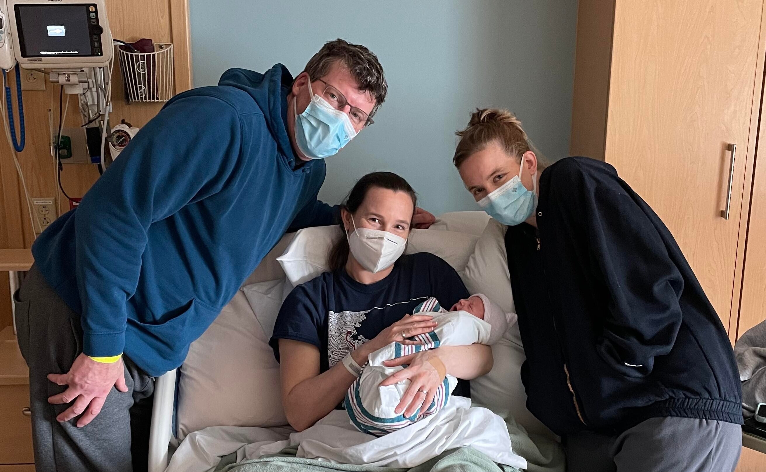 Gestational surrogate in the hospital bed holding the newborn with new parents standing next to her