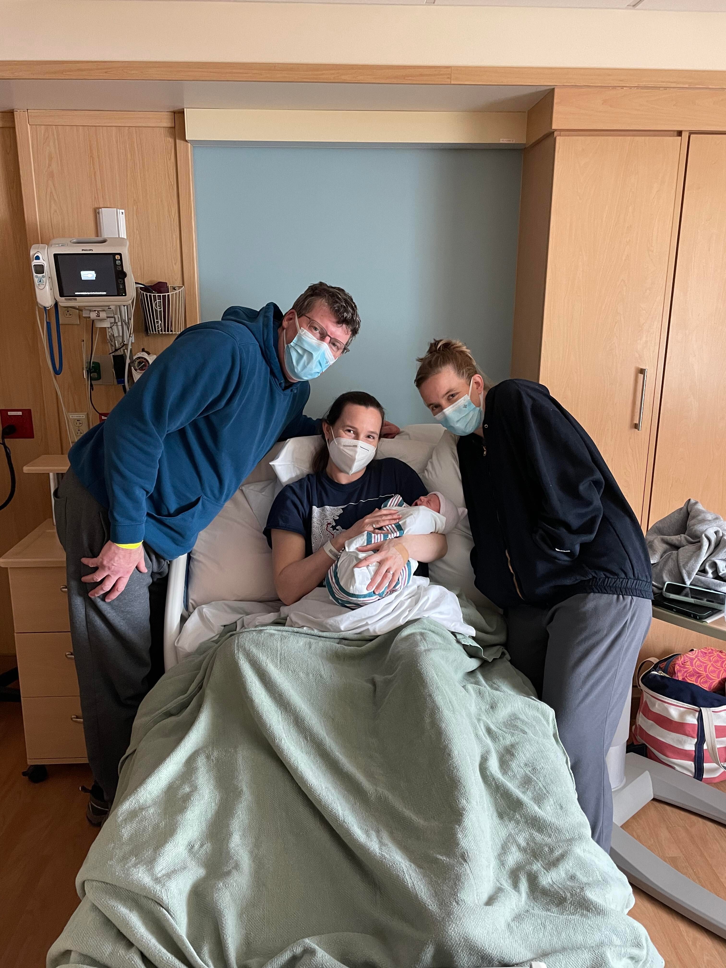 Gestational surrogate in the hospital bed holding the newborn with new parents standing next to her